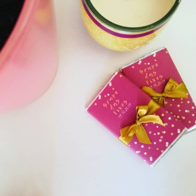 Dois pacotes cor de rosa com laços dourados e texto em amarelo ao lado de vaso rosa e copo decorado
