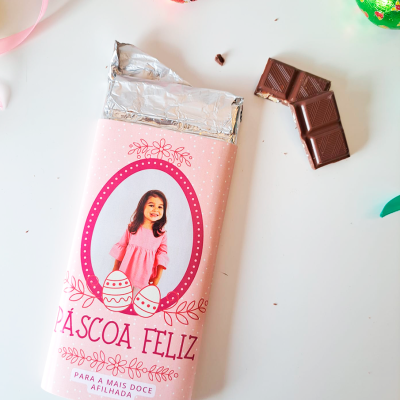 Barra de chocolate com embrulho rosa e texto PÁSCOA FELIZ, ovos de chocolate coloridos e pedaços de chocolate sobre fundo branco