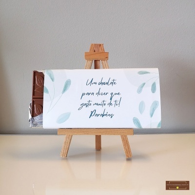 Barra de chocolate embrulhada com mensagem em papel branco com folhas verdes num cavalete de madeira