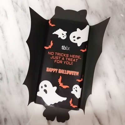 Embalagem preta de Halloween com fantasmas, morcegos e texto em vermelho sobre fundo de mármore