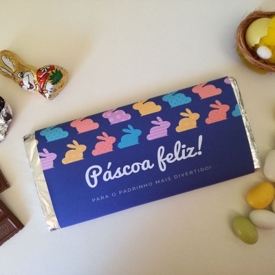 Barra de chocolate com embalagem azul decorada com coelhinhos coloridos e texto de Páscoa rodeada de amêndoas coloridas e enfeites de Páscoa