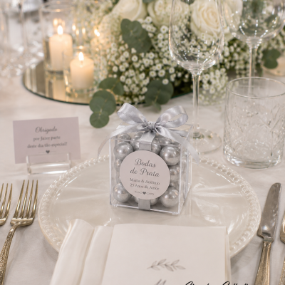 Caixa transparente com bolas prateadas e laço em cima de prato branco numa mesa decorada para celebração.