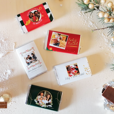 Barras de chocolate com embalagens festivas em vermelho, branco, verde e preto sobre superfície branca com decoração natalina