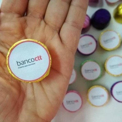 Chocolate redondo com rótulo banco ctt segurado numa mão com vários chocolates semelhantes ao fundo