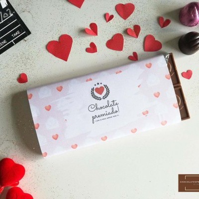 Barra de chocolate com embalagem branca decorada com corações e texto 'Chocolate premiado!' sobre superfície com corações de papel e bombons coloridos