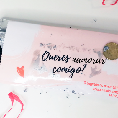Barra de chocolate com embalagem rosa claro e texto romântico, moeda sobre a barra, fundo branco com fitas vermelhas.