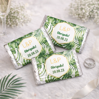 Três chocolates com embalagem de folhas verdes e etiqueta branca com texto L&P Obrigado! 20.08.23 num cenário elegante com alianças e copo de champanhe