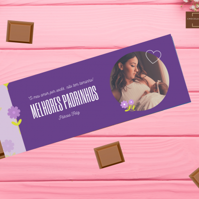 Embalagem de chocolate roxa com frase 'MELHORES PADRINHOS' e imagem de mulher grávida sobre fundo rosa de madeira.