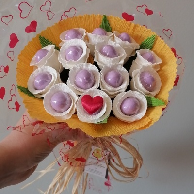 Buquê de cápsulas lilás com decoração em papel amarelo e laço de ráfia, fundo com corações vermelhos.