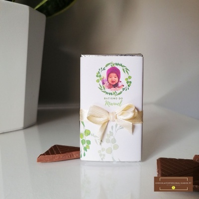 Caixa pequena branca decorada com fotografia de bebé, texto e laço creme, com pedaços de chocolate ao redor