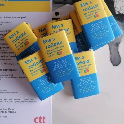 Embalagens azuis e amarelas com mensagem de apoio e bandeiras da Ucrânia e Estónia sobre papel multilingue.