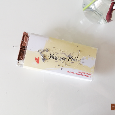 Barra de chocolate com embalagem 'vais ser Pai!' ao lado de um vaso de vidro com flor
