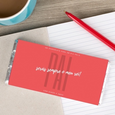 Barra de chocolate com embalagem vermelha e texto, sobre caderno aberto com caneta e chávena.