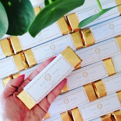 Barra de chocolate com embalagem branca e dourada segurada por mão com várias barras empilhadas ao fundo