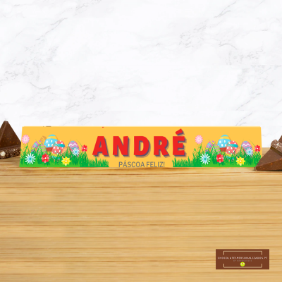 Embalagem amarela retangular com nome ANDRÉ, ovos coloridos e flores sobre mesa de madeira com pedaços de chocolate