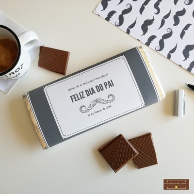 Barra de chocolate com embalagem cinza e branca e texto Feliz Dia do Pai, com pedaços de chocolate e uma chávena de café sobre uma mesa branca