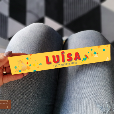 Pulseira amarela de festa com texto LUÍSA FELIZ ANIVERSÁRIO sobre jeans azul claro