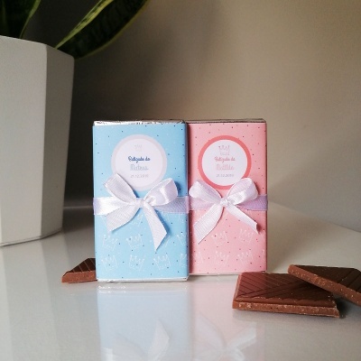 Caixinhas azuis e rosas para lembrança de batizado com laços brancos e etiqueta redonda, ao lado de pedaços de chocolate.