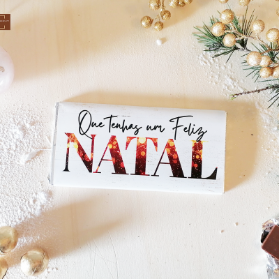 Barra de chocolate com embalagem branca e texto natalício, decoração dourada e galhos verdes com neve artificial.