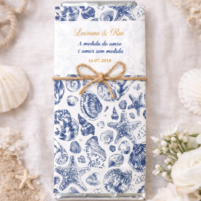 Chocolate embrulhado em papel decorativo com conchas azuis e fio de juta com flores e areia ao redor