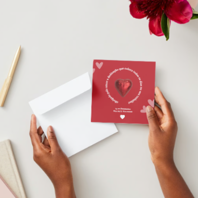 Cartão vermelho com coração brilhante e texto branco em mesa branca com envelope, caneta, cadernos e flor vermelha