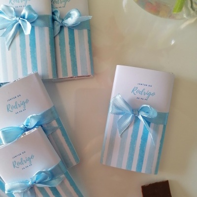 Maio e Junho: dois meses recheados de celebrações! Cinco chocolates embalados com papel branco e riscas azuis com laços azuis e texto 'Jantar do Rodrigo 02.04.23' sobre mesa branca ao lado de pedaços de chocolate e copo com água