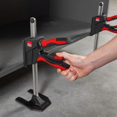 Grampos Bessey vermelhos e pretos segurando prateleira metálica cinza com mão ajustando um deles