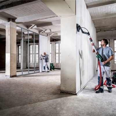 Homem a lixar parede branca com ferramenta elétrica em espaço industrial com chão de cimento