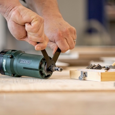 Pessoa a ajustar uma ferramenta elétrica verde numa bancada de madeira com brocas metálicas visíveis