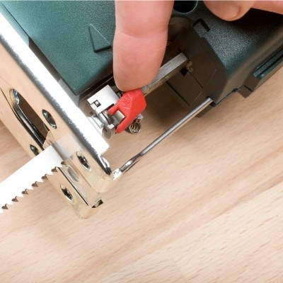 Serra tico-tico elétrica verde escuro com lâmina dentada e botão vermelho sendo ajustado sobre madeira clara