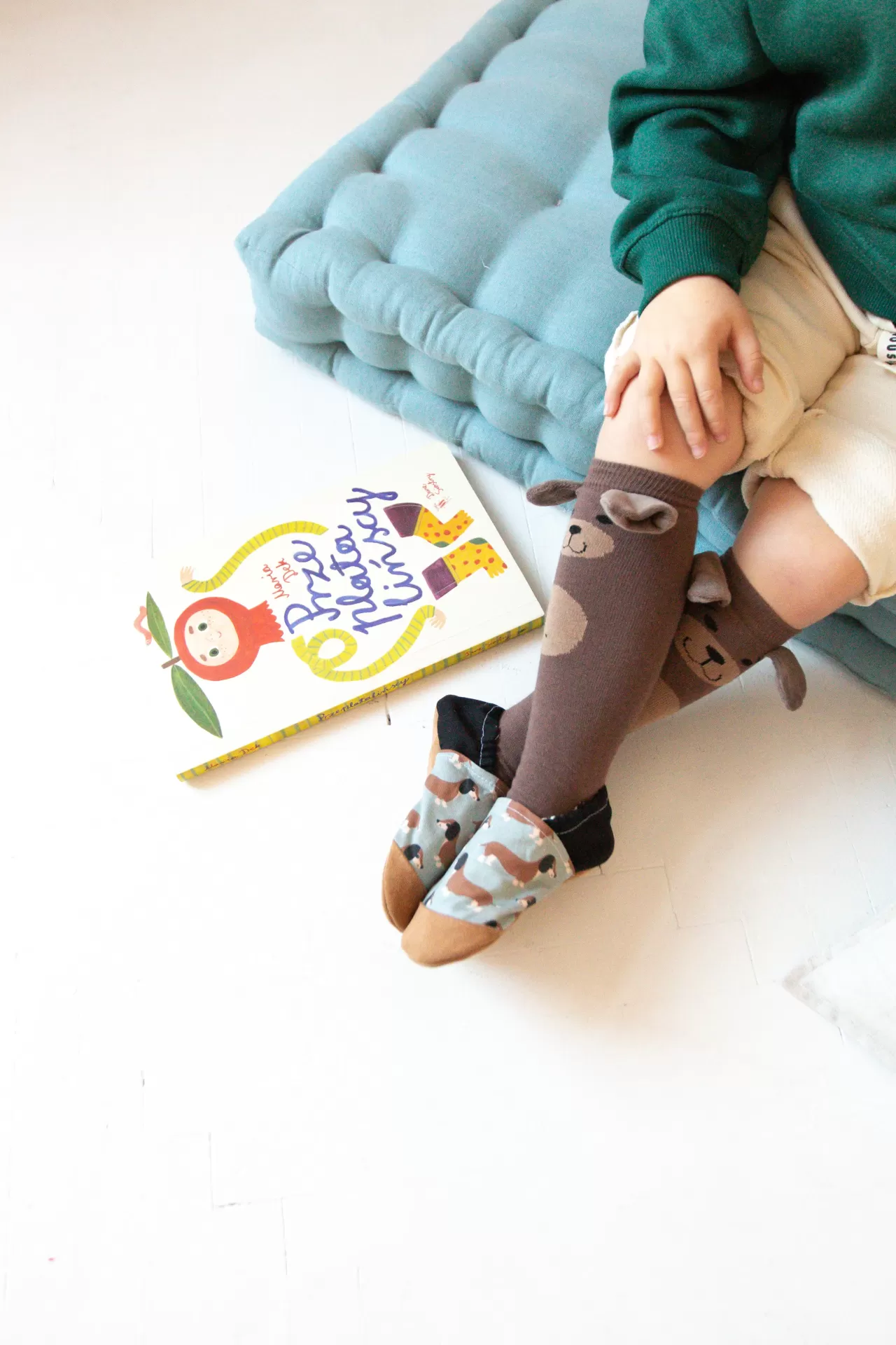 Sapatinhos de Criança em Couro Natural – Cachorro Salsicha Meias de criança castanhas com urso, calçadas, criança sentada em tapete azul e livro colorido ao lado.