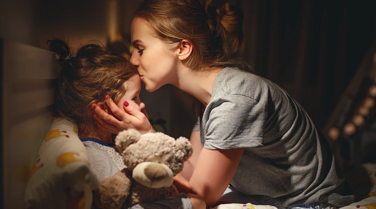Mulher a beijar criança na testa, criança segura urso de peluche na cama