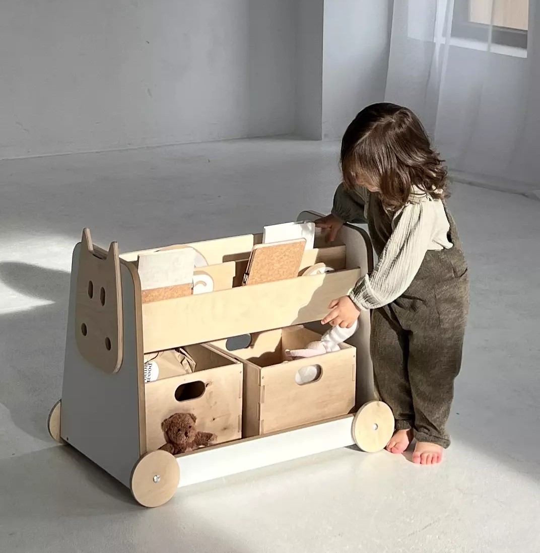 Estante de Madeira Sobre Rodas Carrinho organizador infantil em madeira clara com rodas e gavetas contendo brinquedos e livros, com criança ao lado.