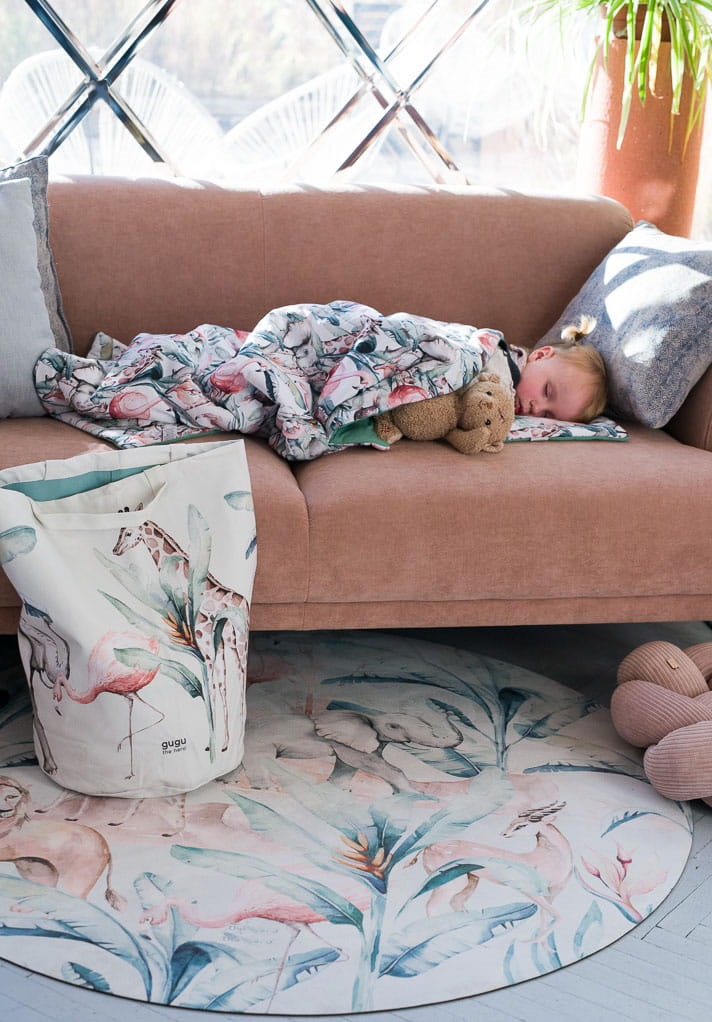 Tapete Redondo ECO Vida Animal Criança a dormir no sofá rosa com manta de flamingos, tapete e cesta com estampa de flamingos.