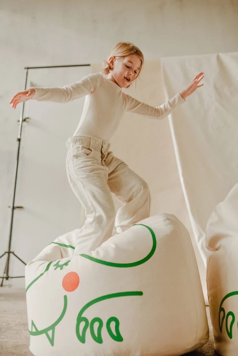 Pufe Bini Grande CROCODILO Menina com roupa clara em cima de puff branco com padrão verde e laranja num espaço interior claro