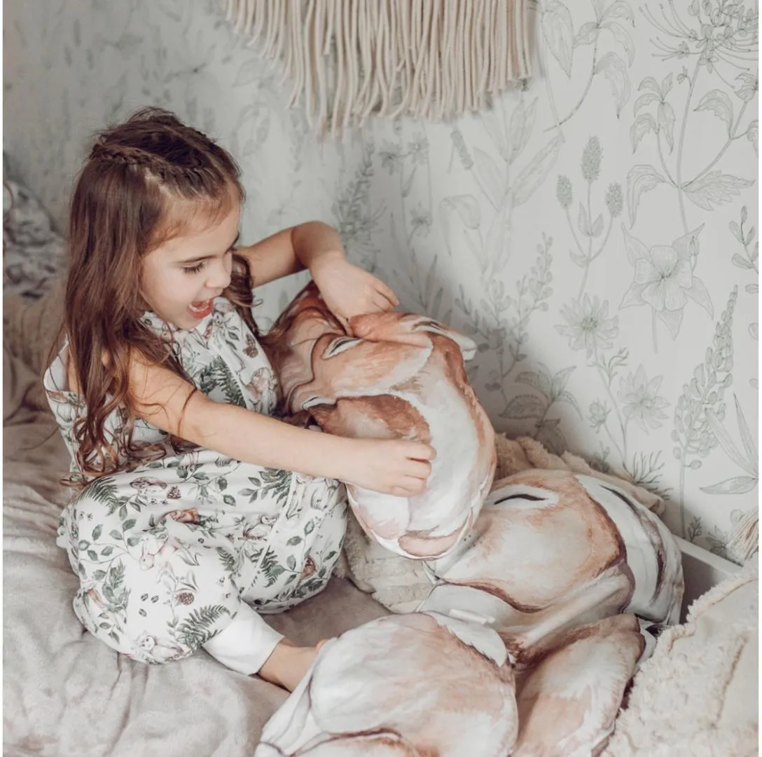 Saco de Dormir com Pernas - Lobos Menina com pijama floral segurando almofada grande com estampa de cervo na cama