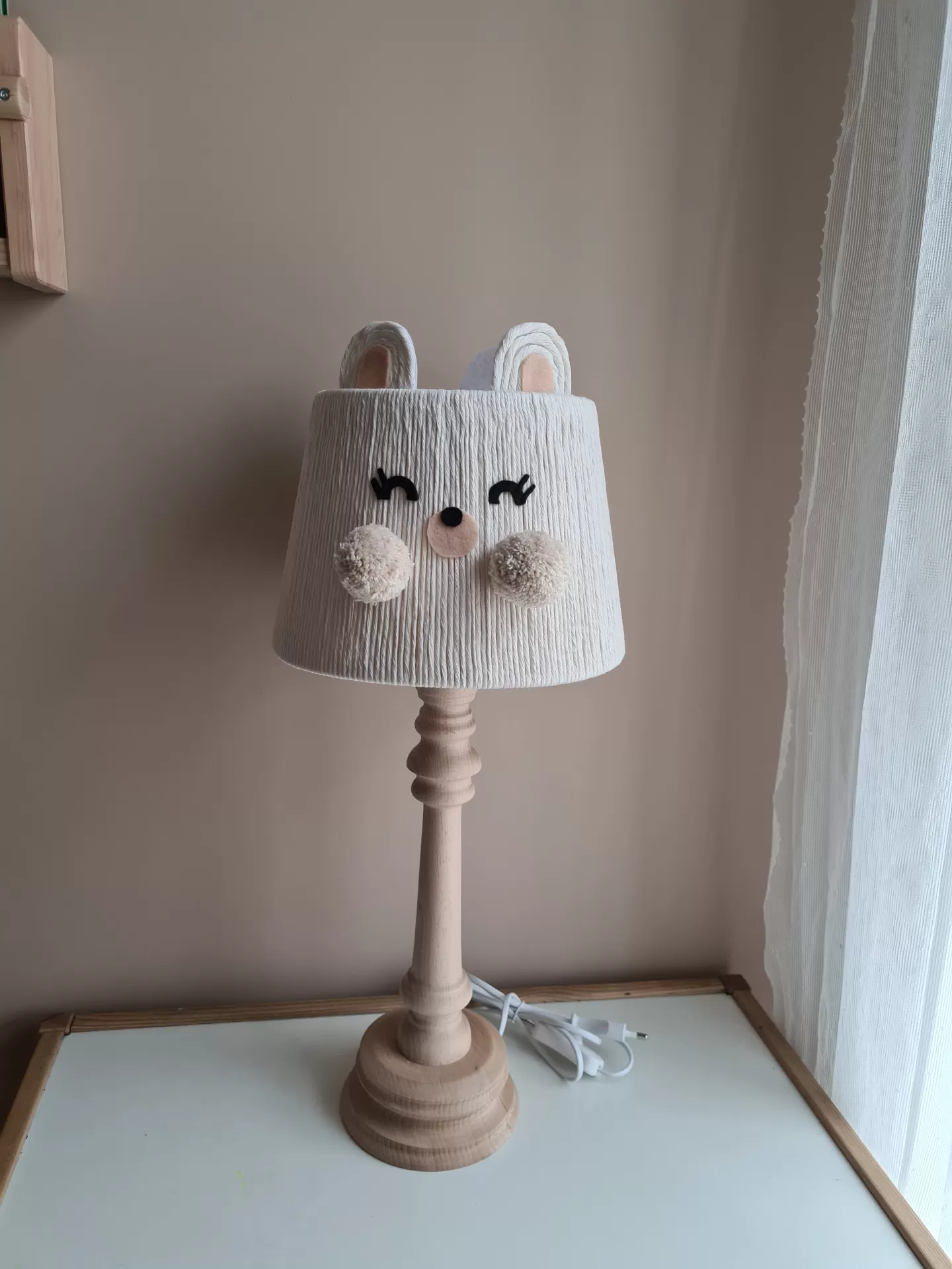 Candeeiro de Mesa URSINHA Candeeiro de mesa infantil com base de madeira e cúpula com carinha desenhada e orelhas, sobre mesa ao lado de cortina branca.