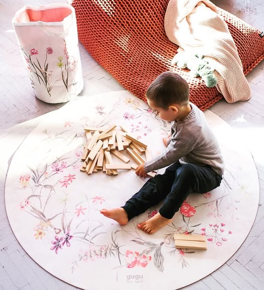 Tapete Redondo ECO de Bebé Jardim de Flores Criança a brincar com blocos de madeira sobre tapete redondo floral, saco de tecido e cobertores ao lado