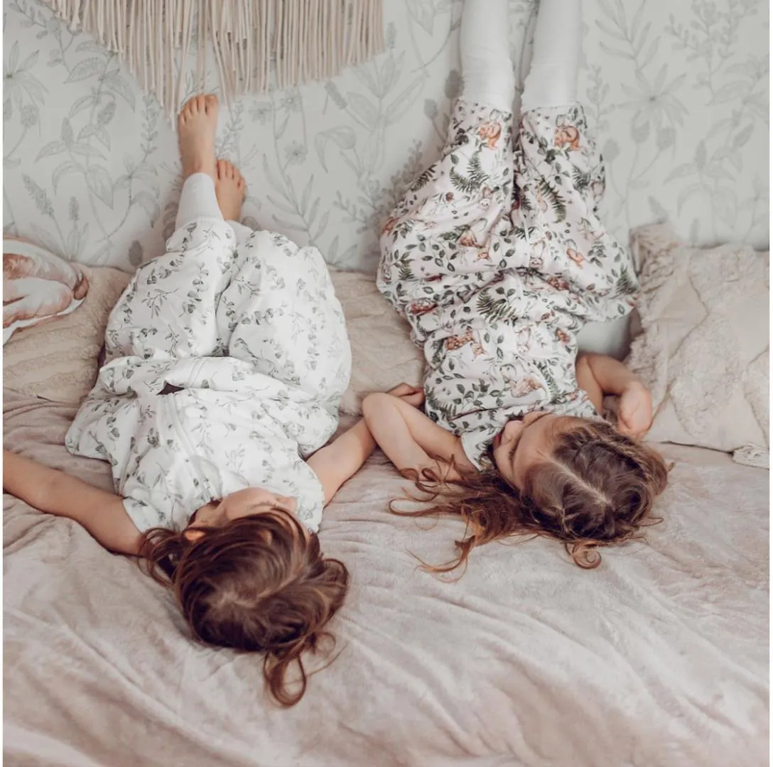 Saco de Dormir com Pernas - Lobos Crianças de pijamas florais deitadas de cabeça para baixo numa cama com almofadas