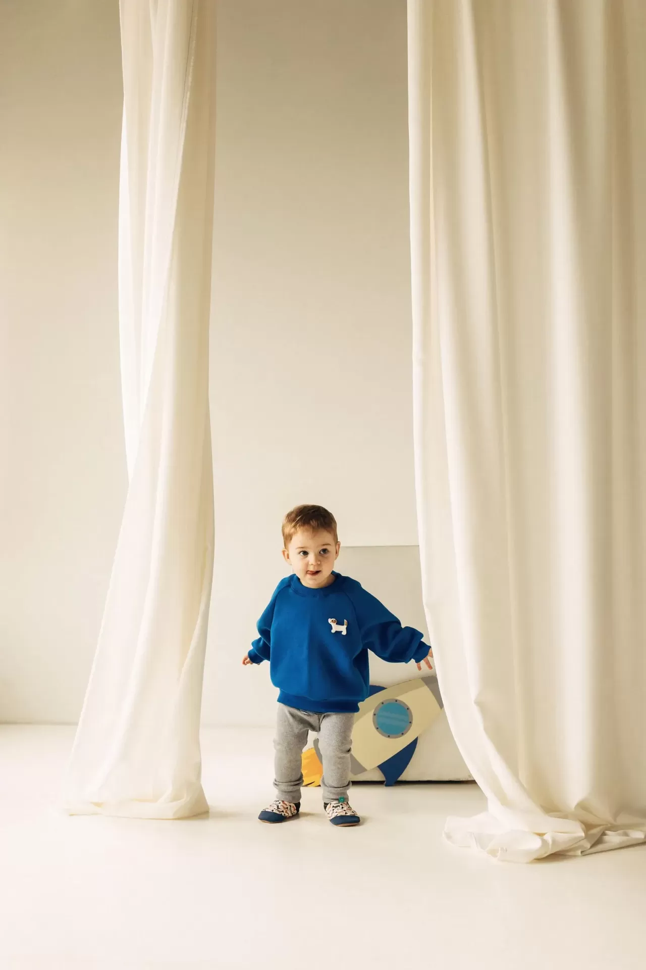 Sapatinhos de Criança em Couro Natural – Cachorros Pequenos Criança com camisola azul e calças cinza em fundo com cortinas brancas e brinquedo foguetão.