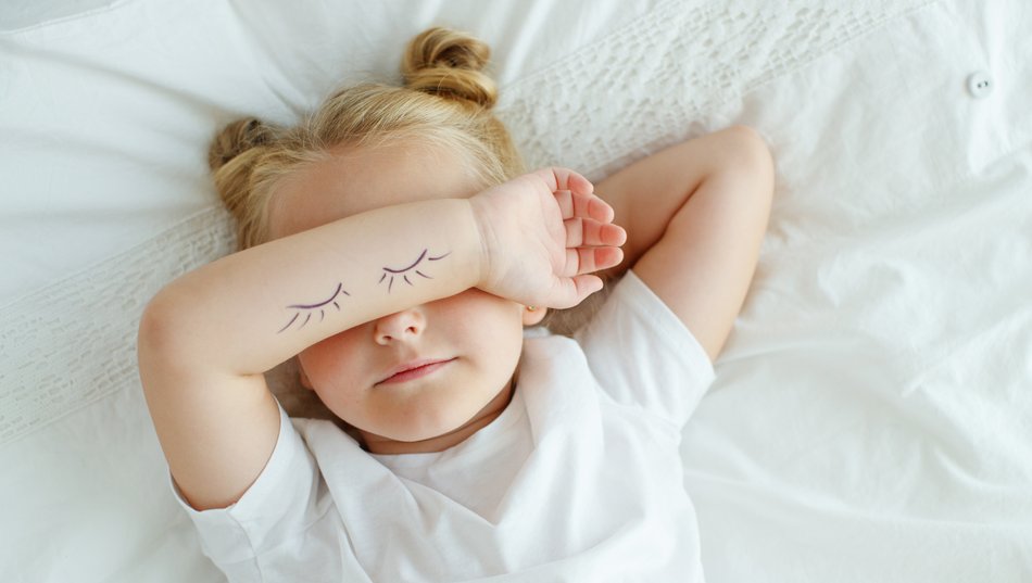 Criança deitada com t-shirt branca e desenhos de cílios no antebraço.