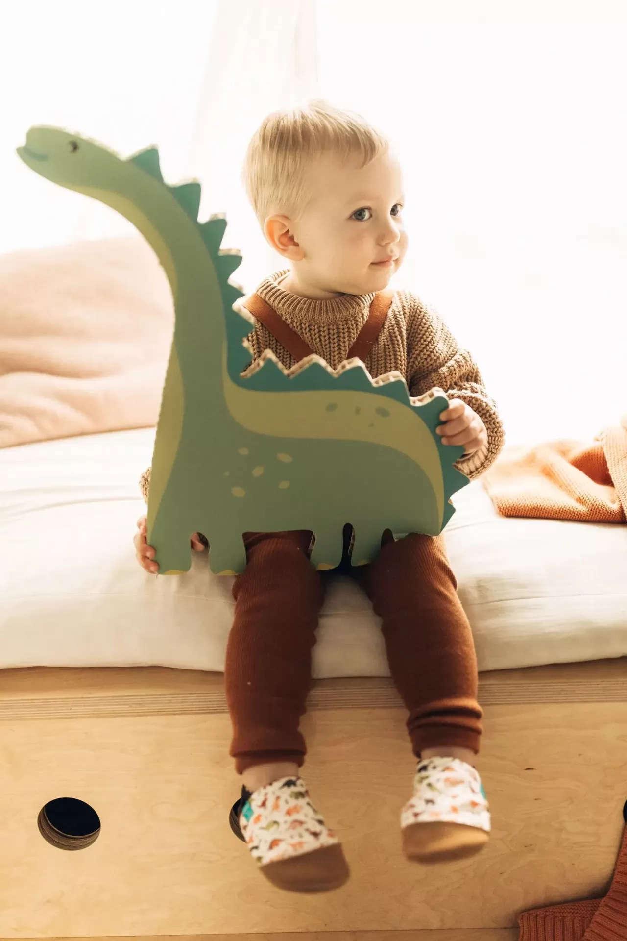 Sapatinhos de Criança em Couro Natural – Dinossauro Marron Criança segurando brinquedo dinossauro verde sentada numa cama de madeira clara
