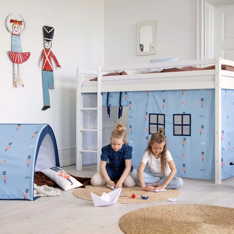 Cama alta branca com cortinas azuis de padrão soldados, crianças a brincar no chão com tenda azul ao lado e bonecos de madeira na parede
