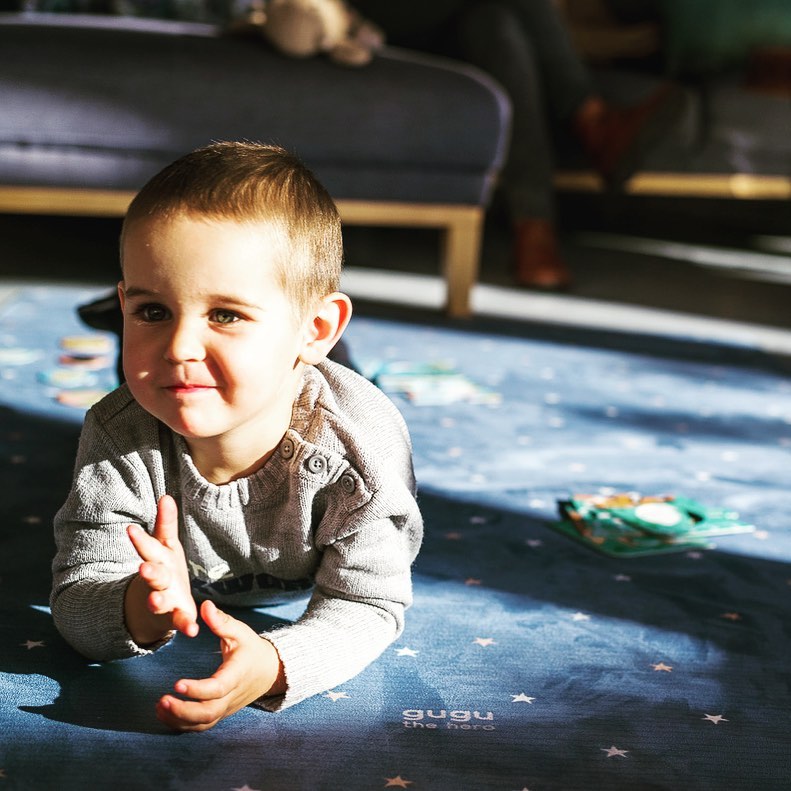 Tapete Grande Borracha Natural GALÁXIA Criança deitada num tapete infantil azul escuro com estrelas brancas e texto 'gugu The Rug'