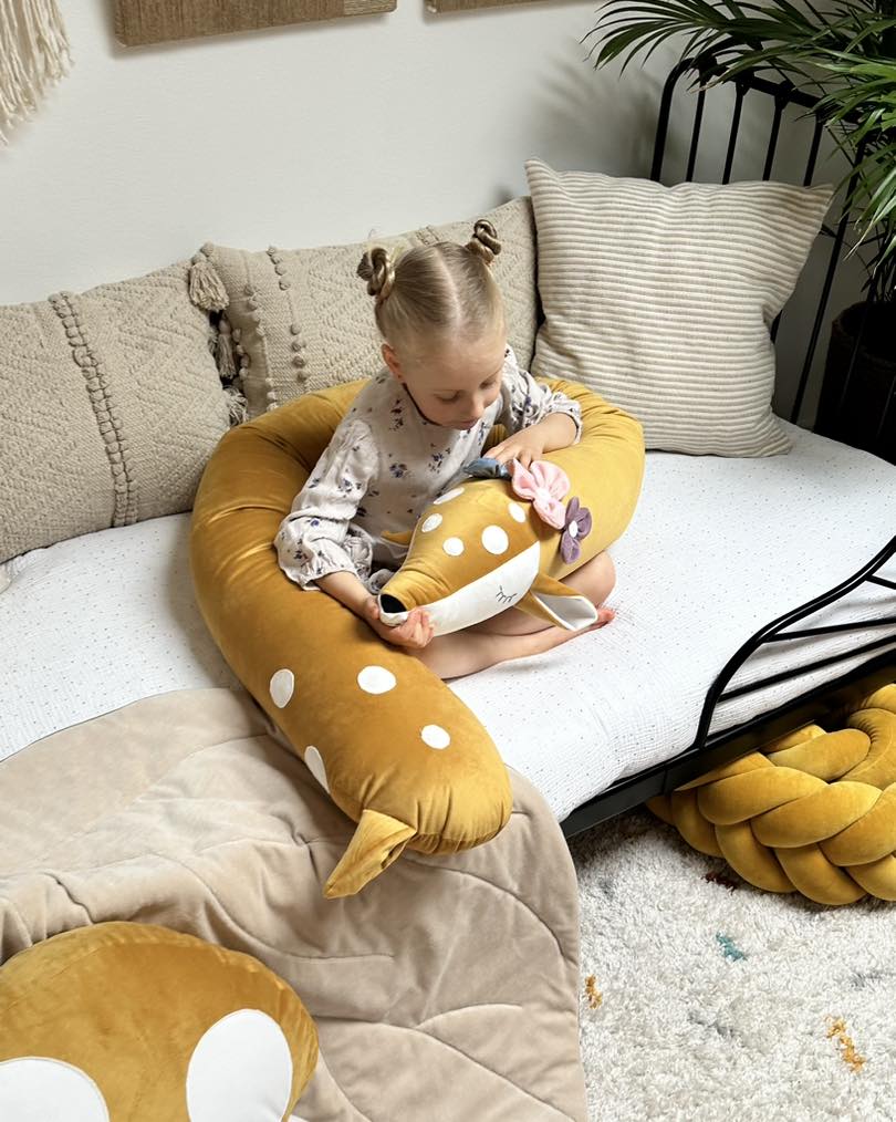 Protetor de Cama Almofada Travesseiro VEADO Criança com almofada em forma de veado em sofá com almofadas neutras num ambiente acolhedor.