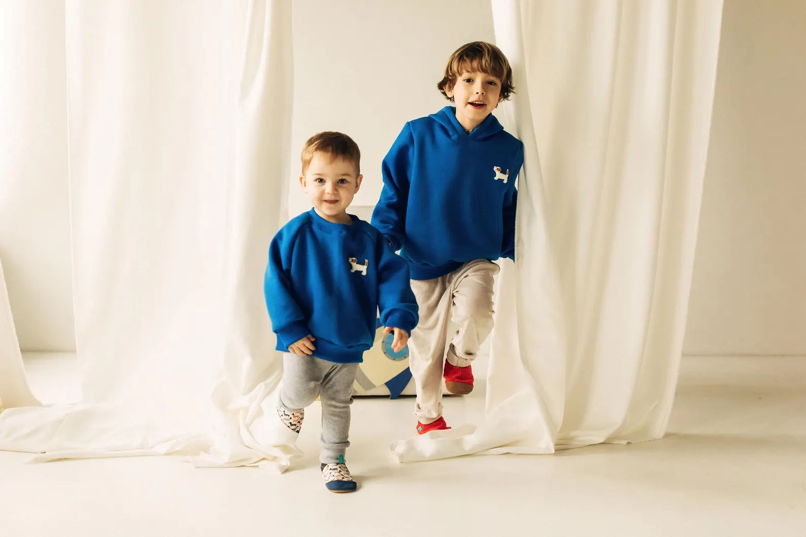 Sapatinhos de Criança em Couro Natural – Cachorros Pequenos Dois rapazes de sweatshirt azul com cão branco, calças claras e ténis no interior com fundo branco.