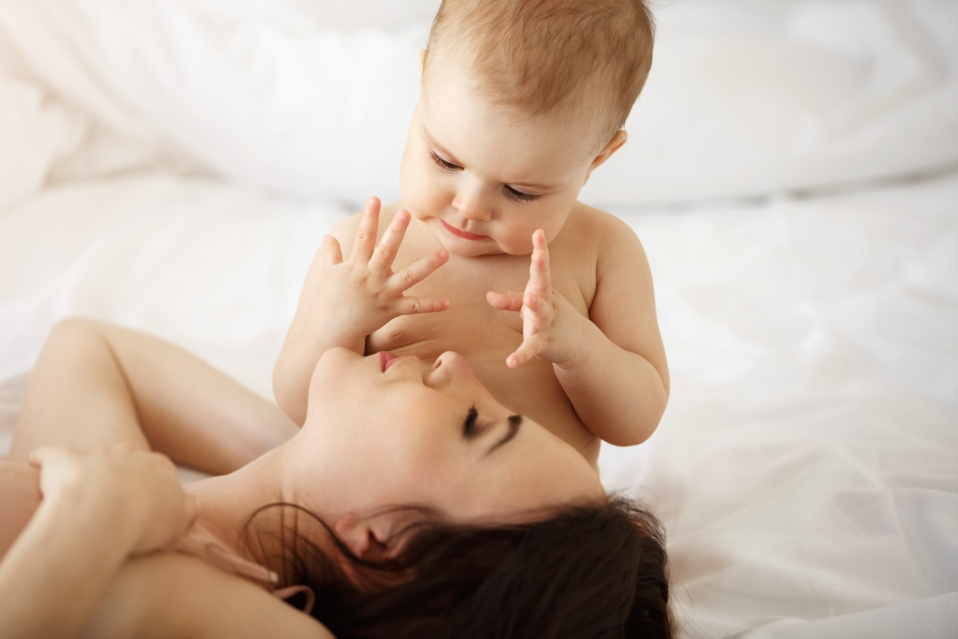 Bebé loiro-avermelhado abraçando mulher numa cama com lençóis brancos