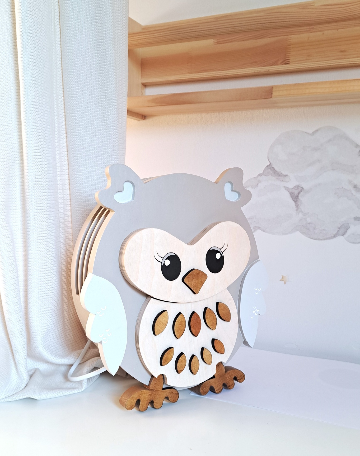 Candeeiro Coruja de Madeira Controlado por SMARTPHONE Abajur coruja de madeira com cores cinza e branco, sobre superfície branca junto a cortina e parede com nuvens