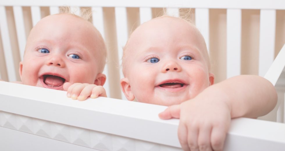 Gémeos: A Magia de Crescerem Juntos Dois bebés sorridentes a segurar a grade branca de um berço