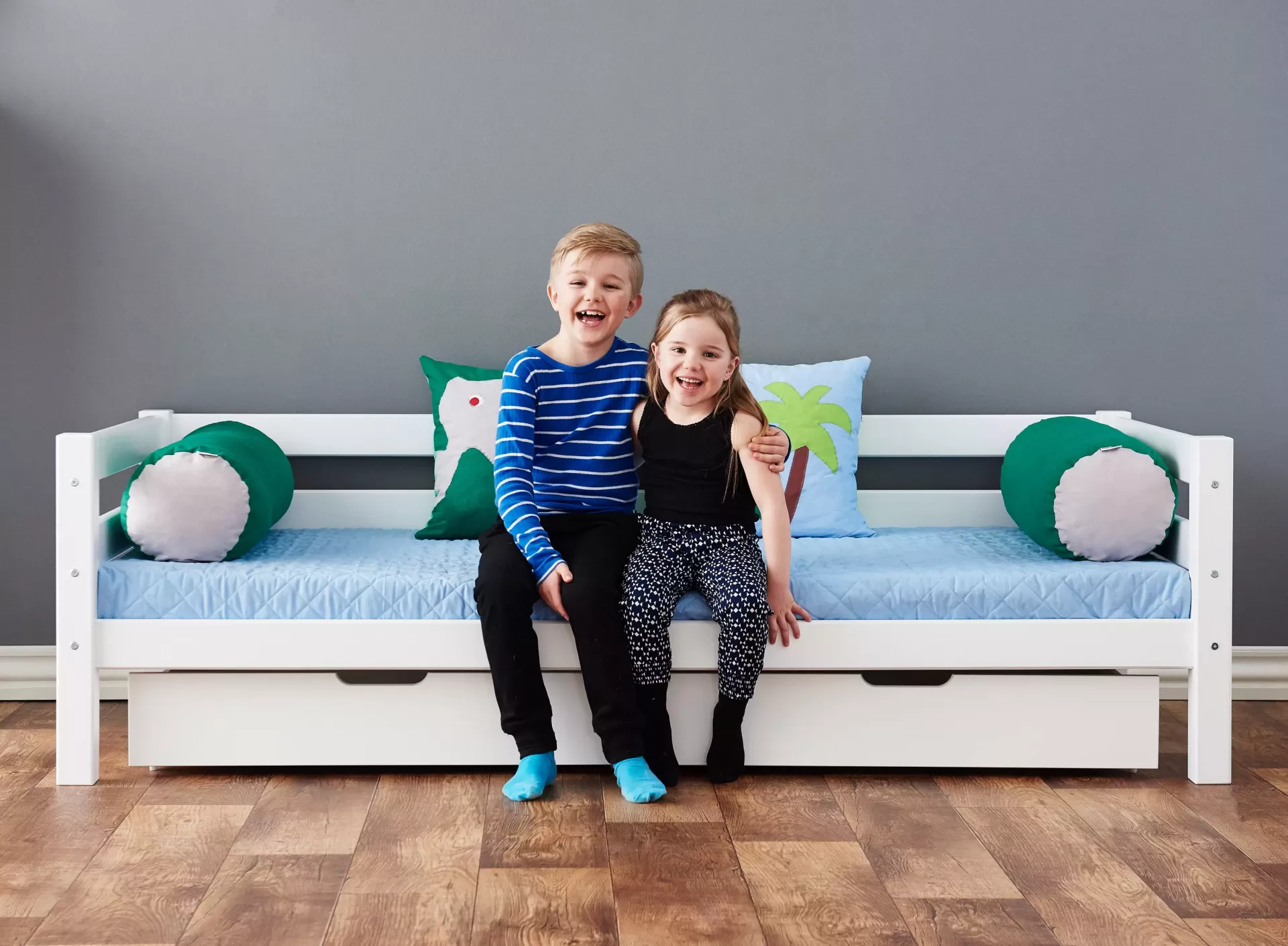 Conjunto de Almofadas Dino Cama branca em madeira com gaveta, colchão azul e almofadas decorativas com crianças sentadas